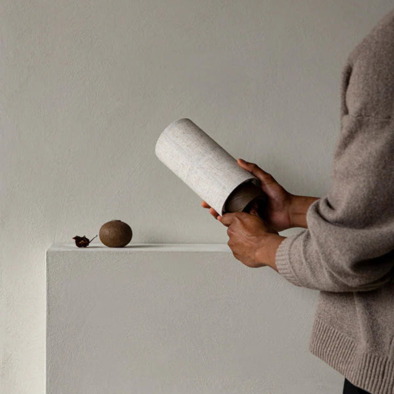Person holding an Audo Copenhagen Hashira Portable Lamp with raw linen shade and dark base near a minimalist mantel, showing its lightweight, rechargeable design in a neutral interior.