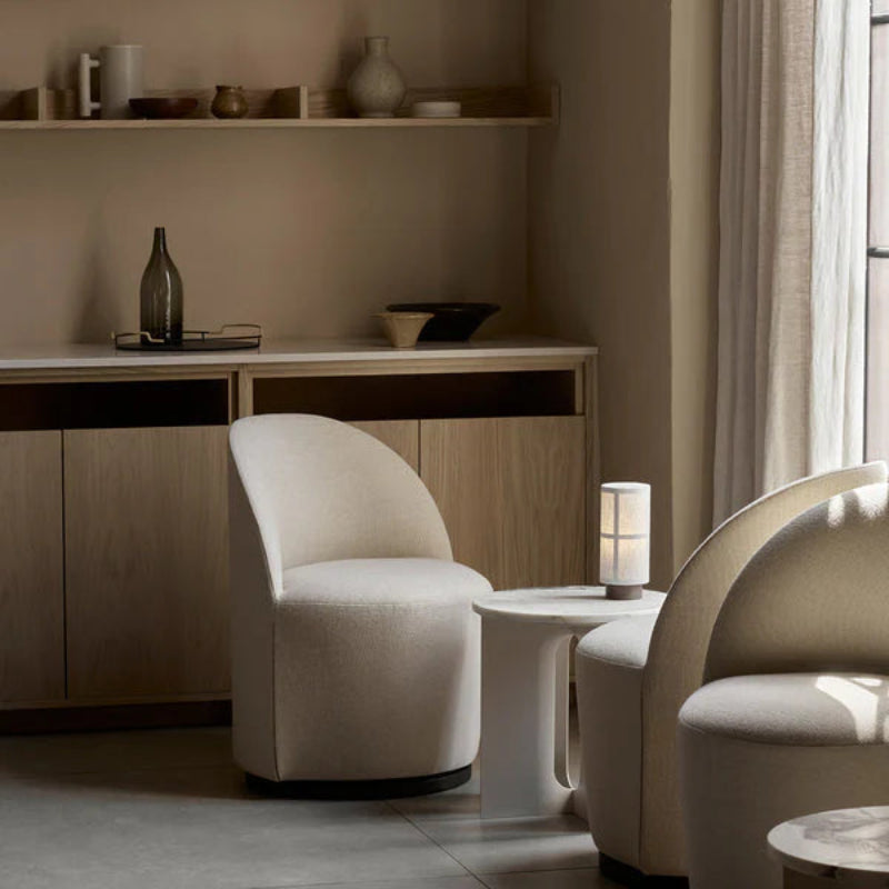 Audo Copenhagen Hashira Portable Lamp with raw linen shade placed on a small white side table between two cream tearoom upholstered chairs in a softly lit living room with neutral shelving and sheer curtains.