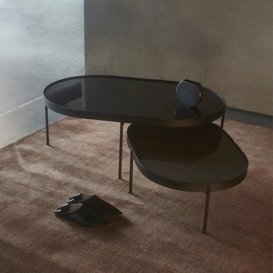Audo Copenhagen NoNo Tables in dark green and brown glass with slender metal frames, styled together on a textured rug in a softly lit modern interior.