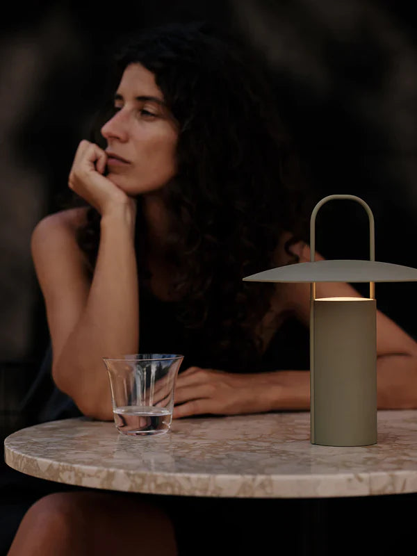 Audo Copenhagen Ray Portable Lamp in olive powder-coated aluminum casting a warm glow on a marble table being used outside with a seated person in soft evening light.