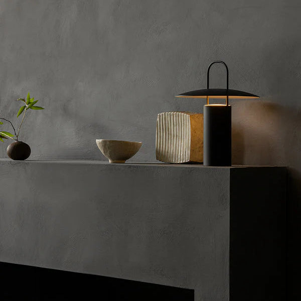 Audo Copenhagen Ray Portable Lamp in black powder-coated aluminum casting a warm glow on a dark textured mantle, styled with sculptural ceramic vessels and a minimalist branch.