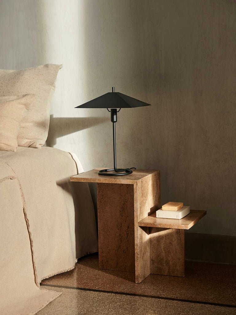 Ferm Living Distinct Side Table in Dark Brown Travertine styled as a bedside table with a black lamp and stacked books in a warm, minimalist bedroom.
