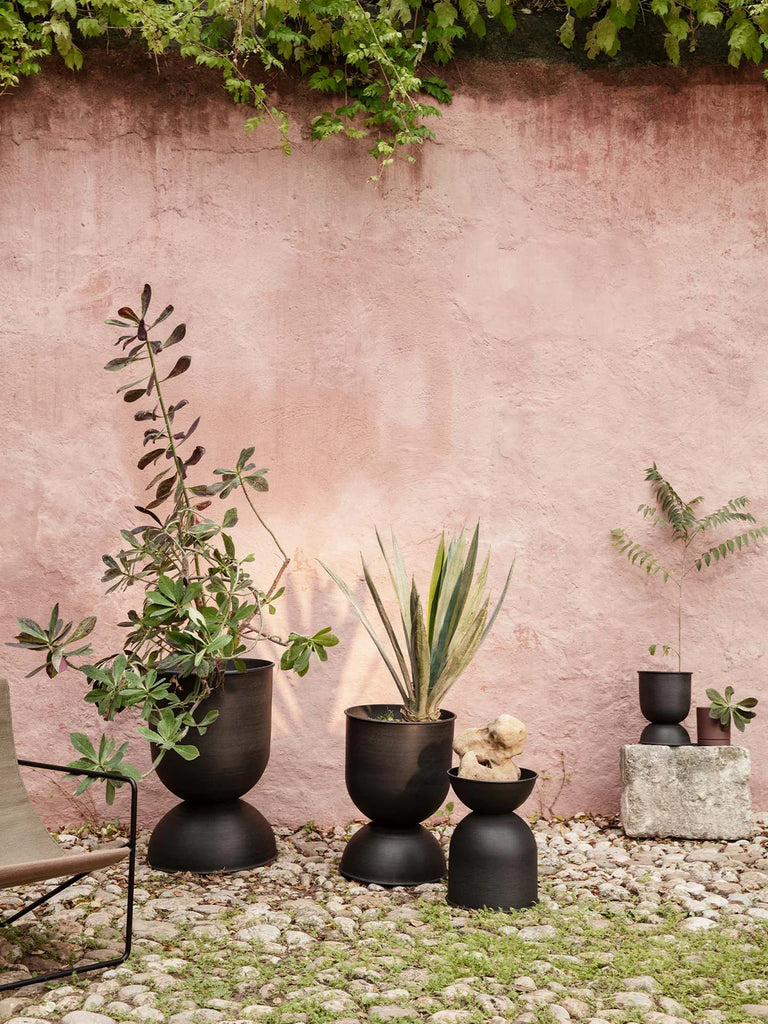 Ferm Living Hourglass Pots in Black displayed outdoors against a pink stucco wall, filled with sculptural greenery and arranged on a cobblestone patio.