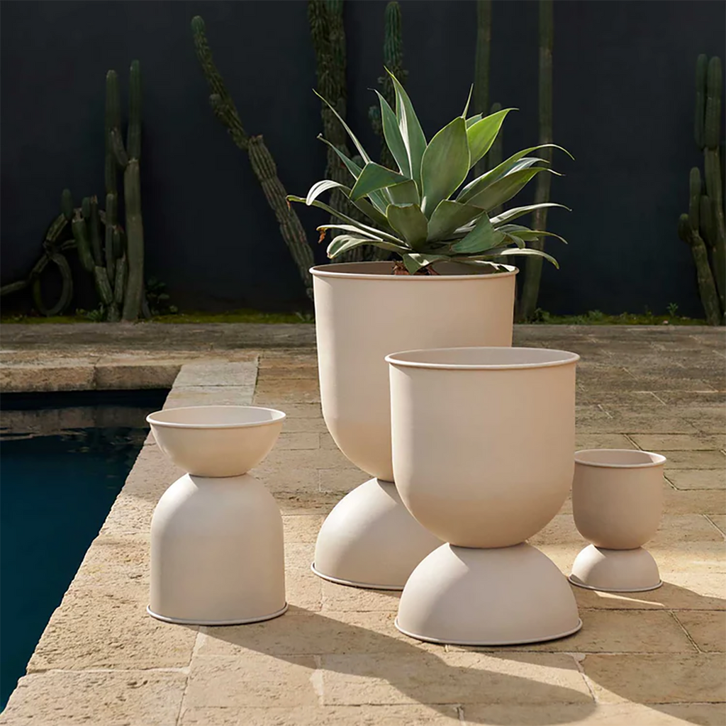 Ferm Living Hourglass Pots in Cashmere powder-coated iron in varying sizes, styled outdoors beside a pool with cacti and agave plants against a dark stucco wall.