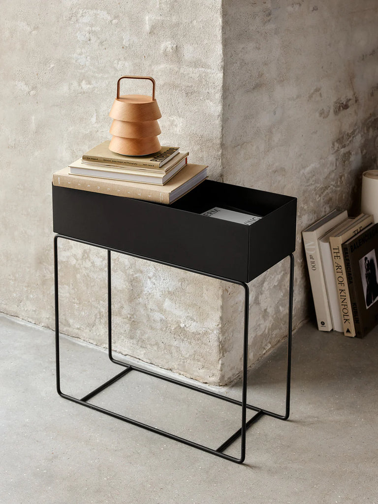 Ferm Living Plant Box in Black styled against a textured concrete wall, used as modern storage with stacked books and a sculptural wooden lamp