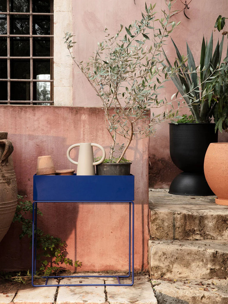 Ferm Living Plant Box in Bright Blue styled outdoors against a pink stucco wall with terracotta pots, olive tree, and sculptural greenery.