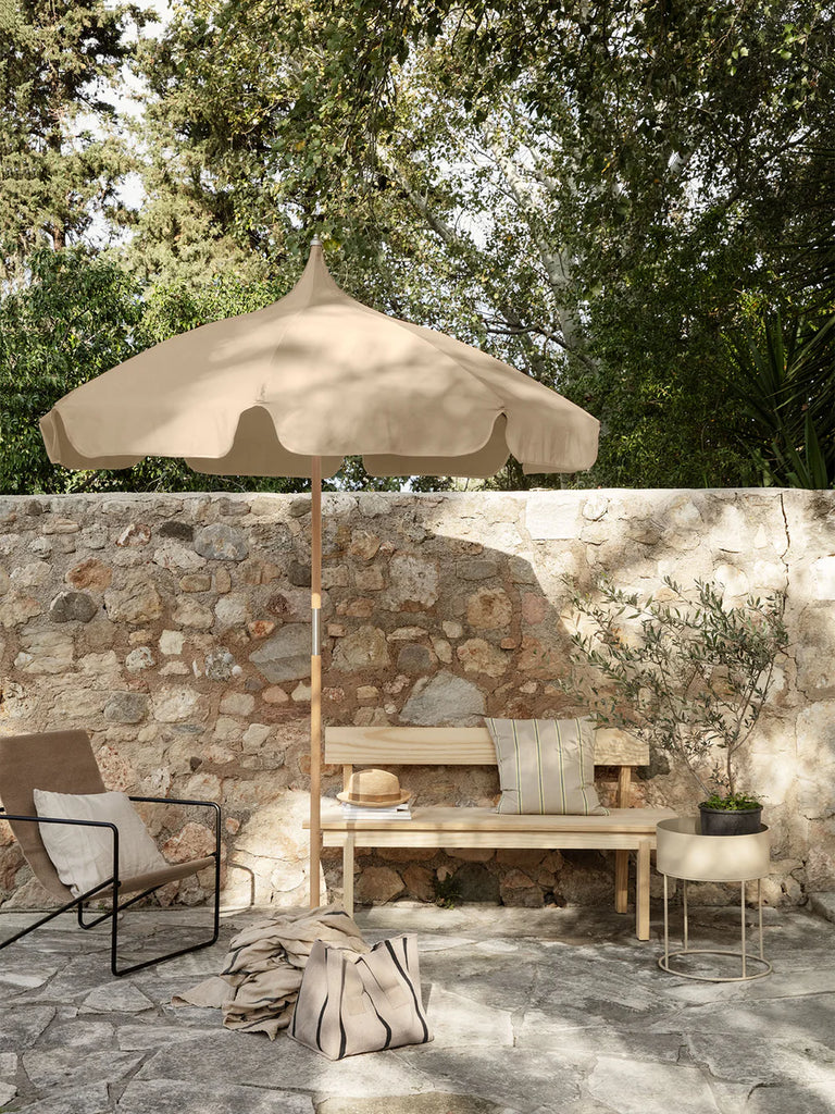 Ferm Living Plant Box Round in Light Grey styled on a stone patio with wooden bench, umbrella, and greenery in a serene outdoor setting.