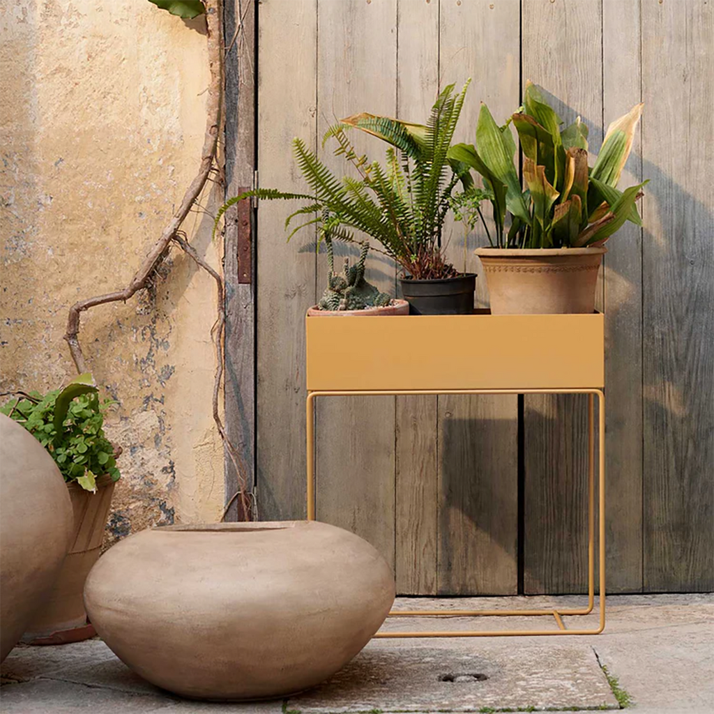Ferm Living Plant Box in Straw styled outdoors against a rustic wooden gate with potted plants and trailing greenery in warm natural light
