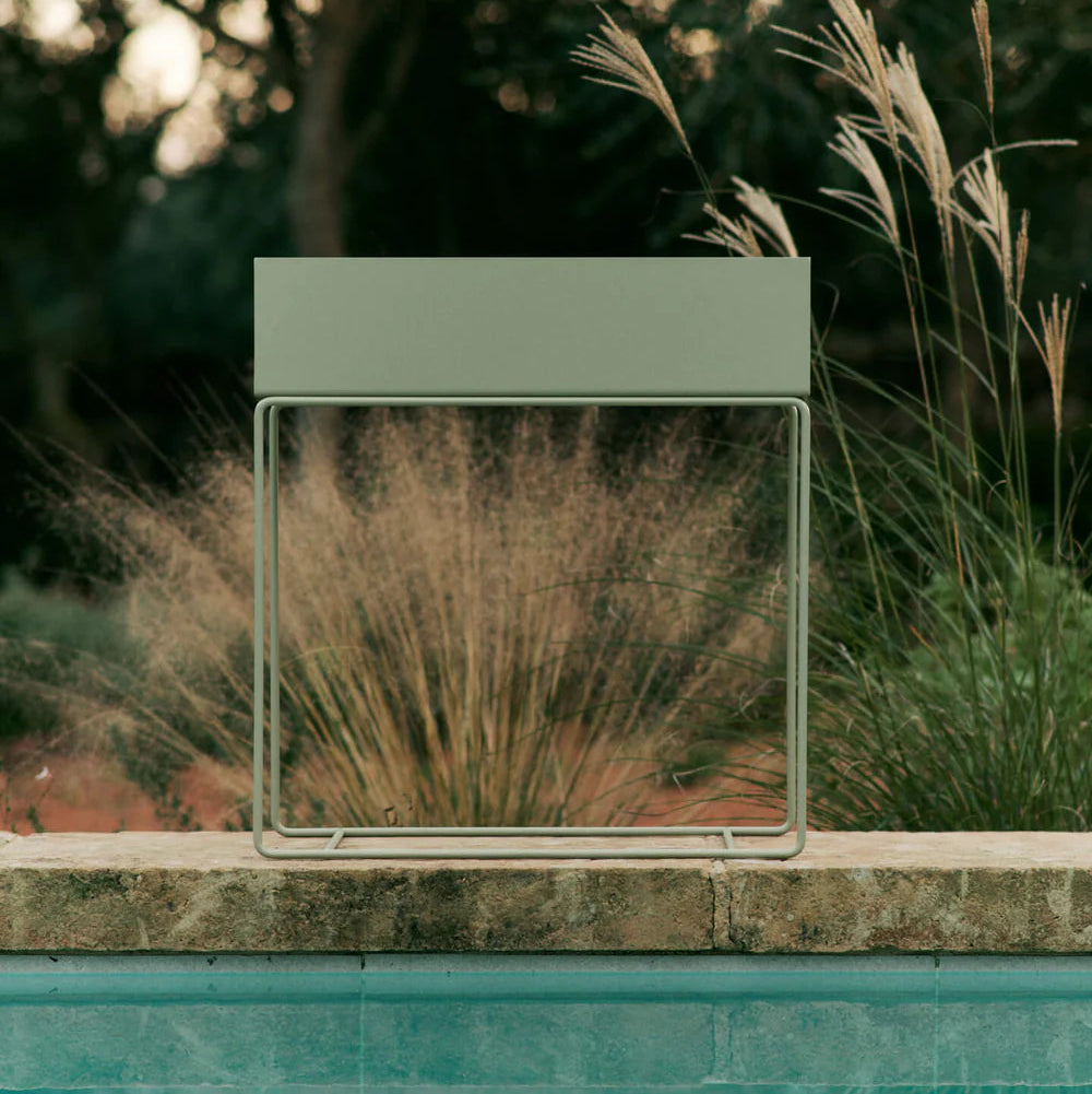 Ferm Living Plant Box in Tea Green placed beside a pool, surrounded by tall ornamental grasses and lush greenery in a serene outdoor setting