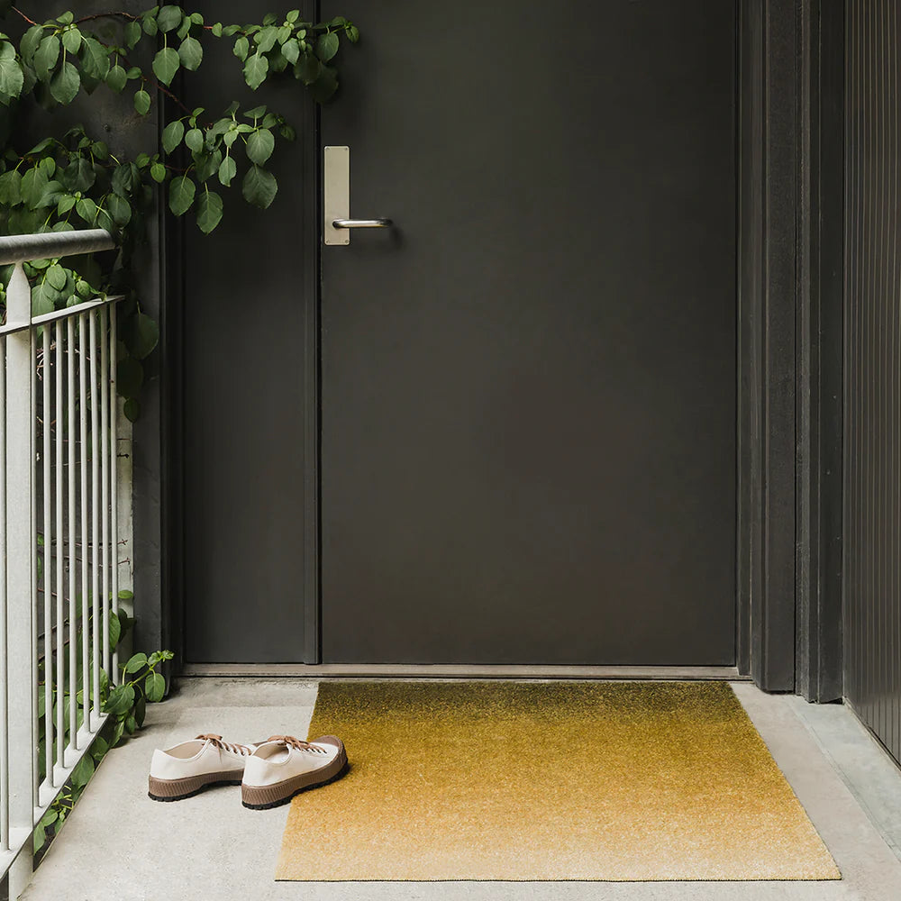 Heymat Dis Moss ombré doormat, styled as an outdoor mat against black exterior door with bright green plants.