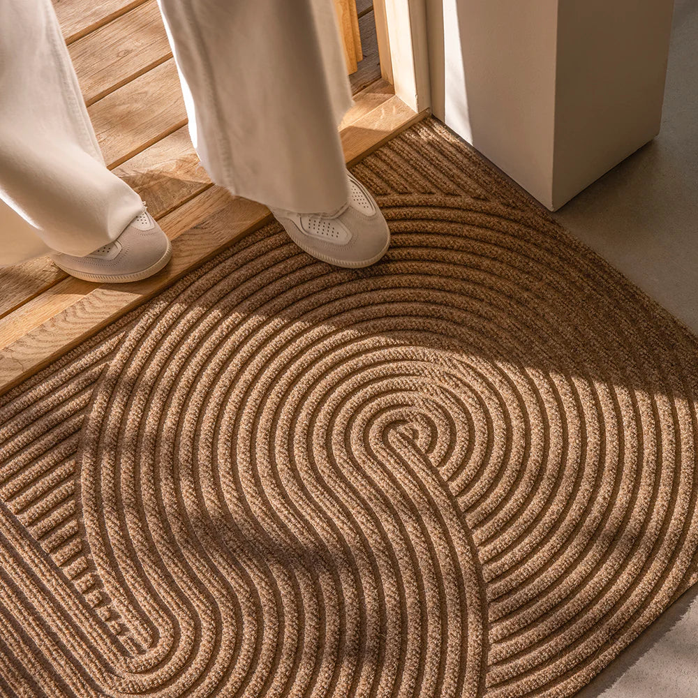 Heymat Sand doormat with Zen garden-inspired 3D ripple pattern, styled in modern Scandinavian entryway with cement floors and person for refrence.