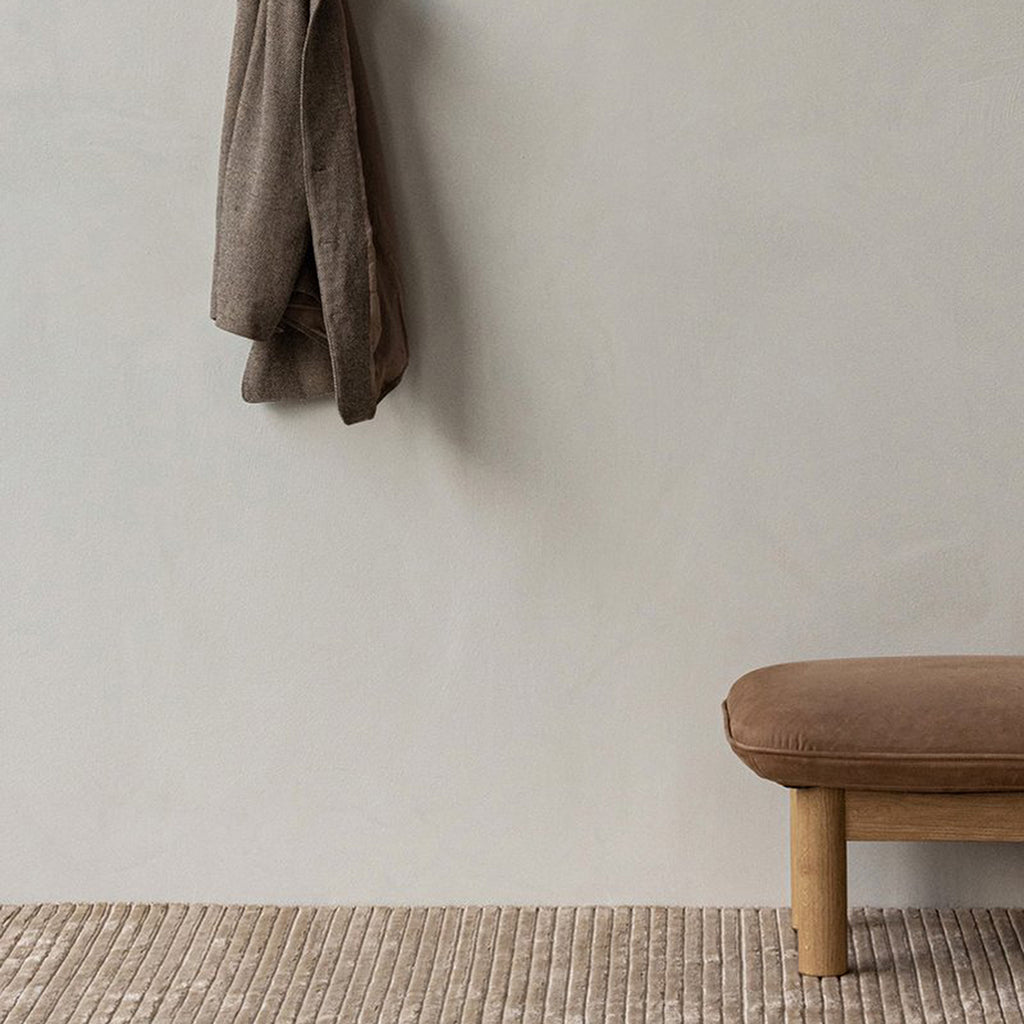 Audo Copenhagen Brasilia Ottoman in natural oak with Dunes Camel leather upholstery, styled in a neutral interior beside a textured rug and hanging coat.