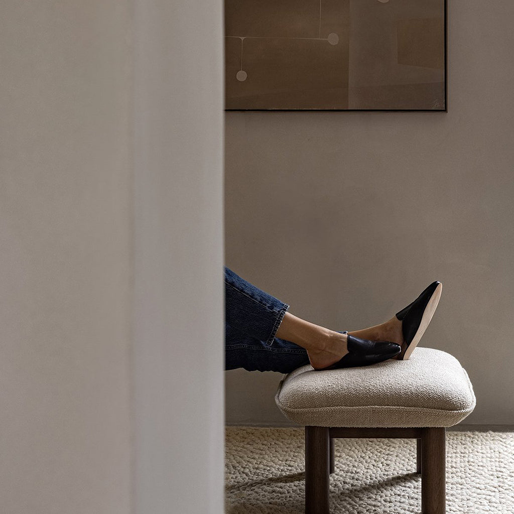 Audo Copenhagen Brasilia Ottoman in dark stained oak with 02 Menu Bouclé upholstery, styled in a minimalist living space with a person resting their feet on the cushioned seat.