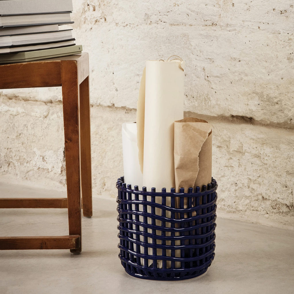 Ferm Living Ceramic Basket Large in Blue — glossy hand-glazed ceramic basket styled in a modern interior beside a wooden table, holding rolled paper and craft materials against a textured stone wall