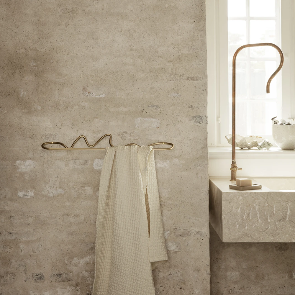 Curvature Towel Hanger in Brass by Ferm Living mounted on wall holding linen towel — sculptural handcrafted brass design.