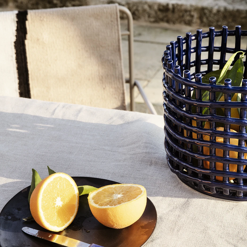 Ferm Living Ceramic Basket Large in Blue — glossy hand-glazed ceramic basket styled outdoors on a linen-draped table beside a sliced orange, bathed in natural light