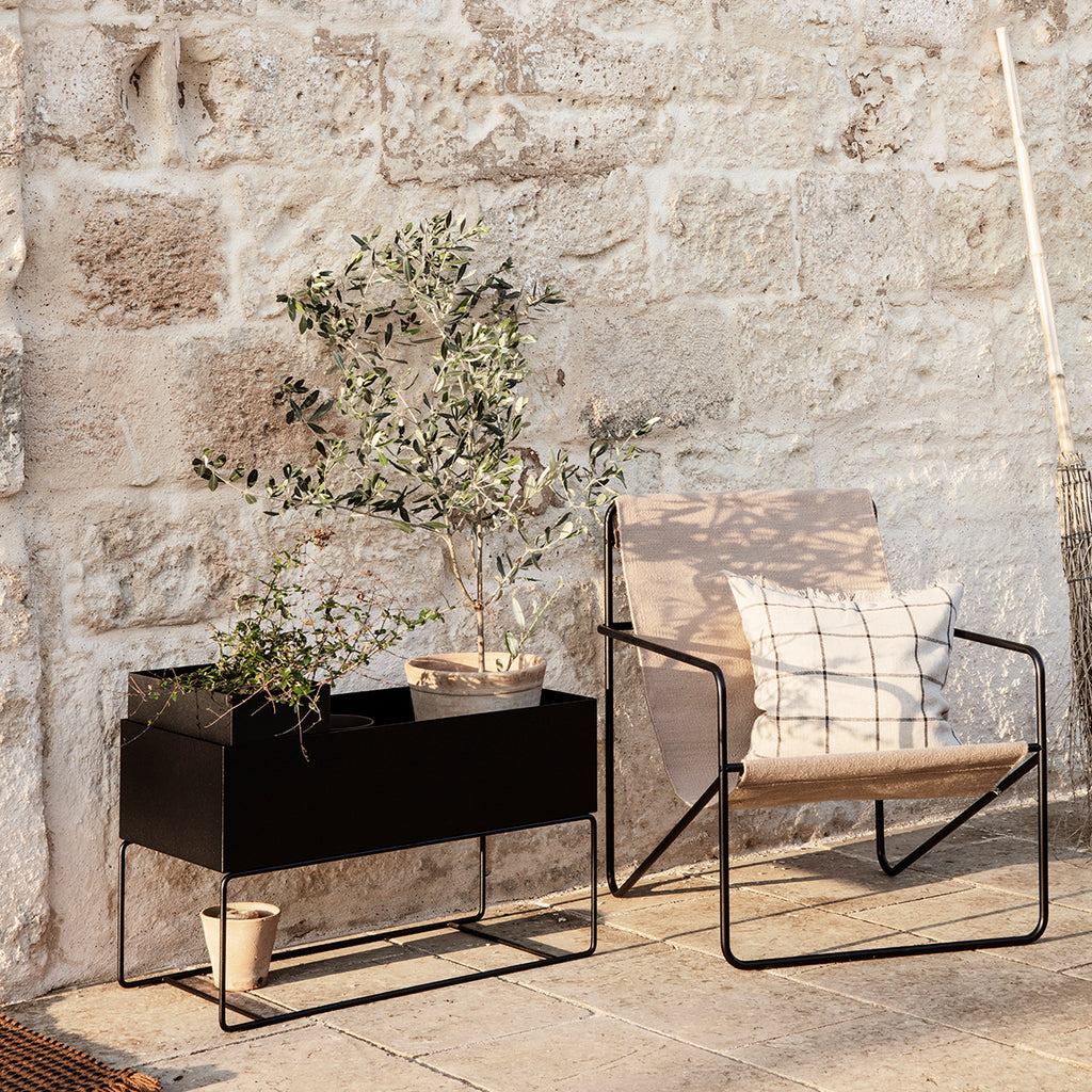 Ferm Living Large Plant Box in black powder-coated steel styled outdoors with potted plants beside a modern lounge chair, creating a relaxed, sunlit patio setting against a textured stone wall.