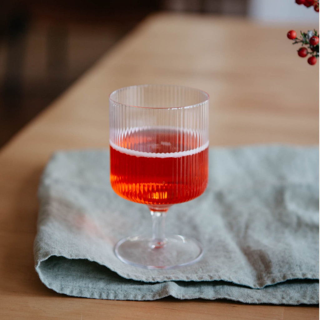 Ferm Living Ripple Wine Glass in Clear filled with red cocktail, placed on a soft green linen napkin atop a wooden table.