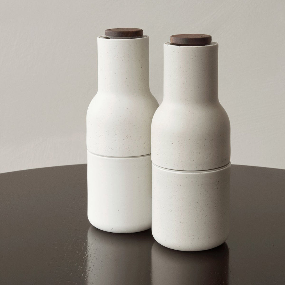 Audo Copenhagen Bottle Grinders in sand with walnut tops, ceramic-look finish, minimalist Scandinavian salt and pepper mills on a dark table