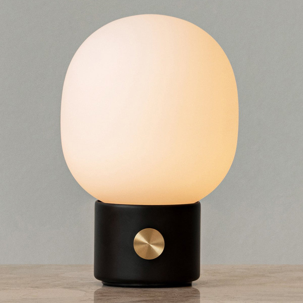 Audo Copenhagen JWDA Portable Lamp in black with illuminated frosted glass shade and brass dimmer, shown on a neutral background sitting on marble table.