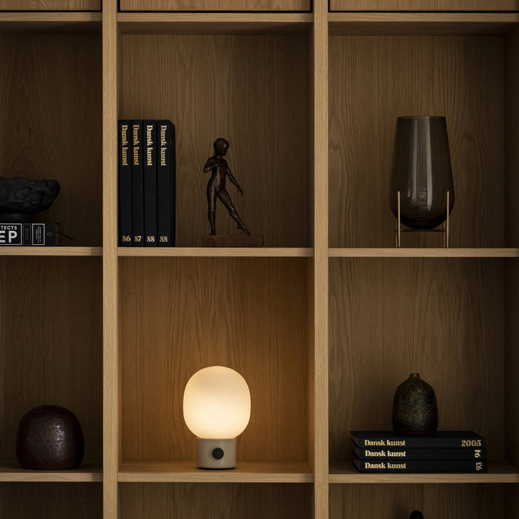 Audo Copenhagen JWDA Portable Lamp in Alabaster White with frosted glass shade casting a warm glow on a wooden bookshelf, surrounded by sculptural decor and books.
