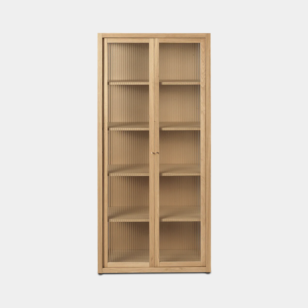 Studio shot of the Ferm Living Reed Vitrine in natural oak with reeded glass doors and brass handles.